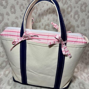 May include: A cream-coloured canvas tote bag featuring dark navy blue vertical straps and handles. The top section is accented with a pink and white striped fabric panel, finished with two small decorative bows tied on the front handles.