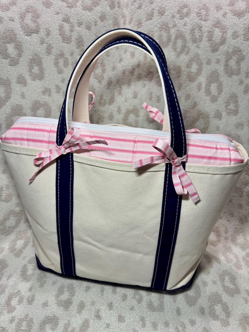 May include: A cream-coloured canvas tote bag featuring dark navy blue vertical straps and handles. The top section is accented with a pink and white striped fabric panel, finished with two small decorative bows tied on the front handles.