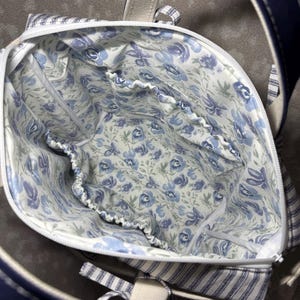 May include: A fabric tote bag with a white zip closure and a dark blue exterior. The interior is lined with a white fabric featuring a blue floral pattern and includes multiple elasticated pockets along the inner perimeter for organisation.