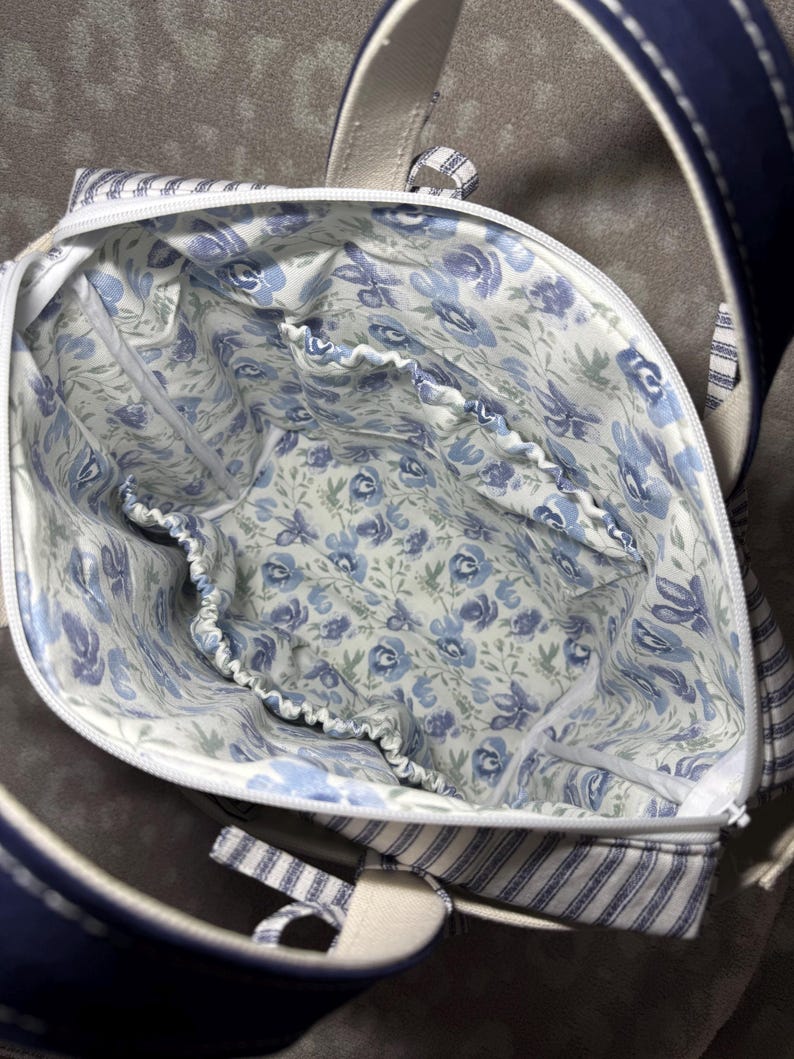 May include: A fabric tote bag with a white zip closure and a dark blue exterior. The interior is lined with a white fabric featuring a blue floral pattern and includes multiple elasticated pockets along the inner perimeter for organisation.