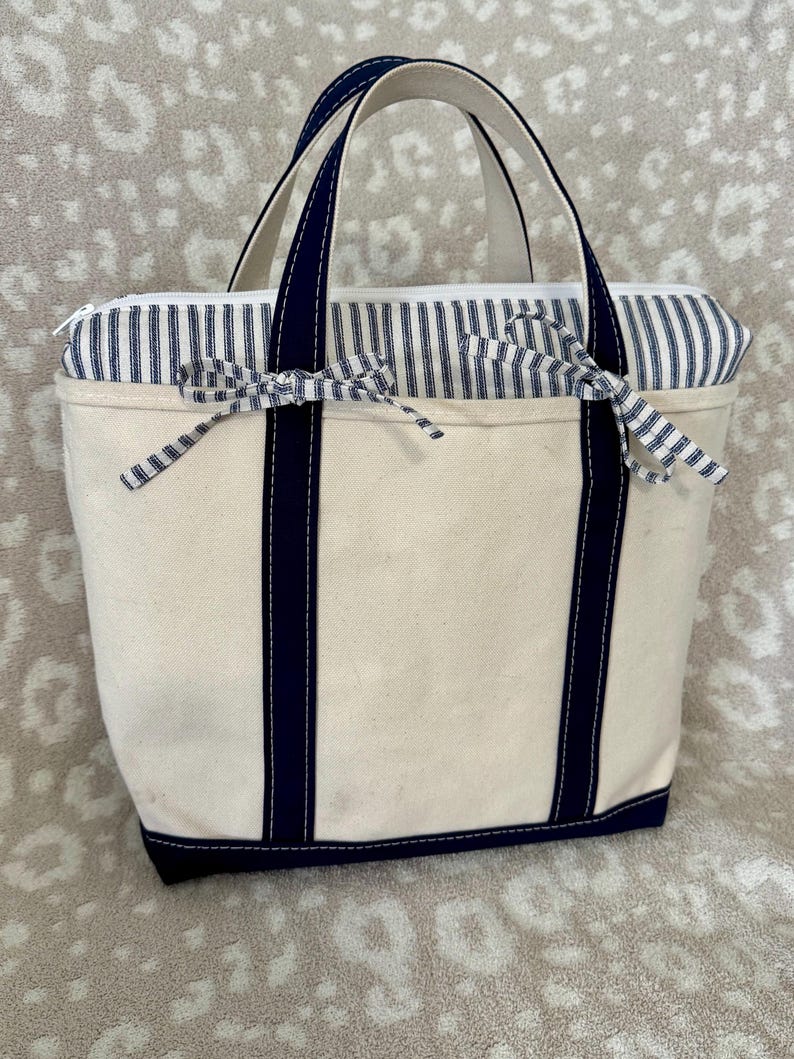 May include: A cream-coloured canvas tote bag featuring dark navy blue vertical straps and a matching base. The upper section has a blue and white pinstriped fabric panel with two decorative fabric bows tied at the front and a white zip closure.