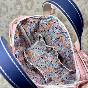 May include: A fabric bag with a pink and white striped exterior and a navy blue handle featuring white stitching. The interior is lined with a floral pattern in shades of pink, orange, blue, and green, and includes an elasticated pocket.