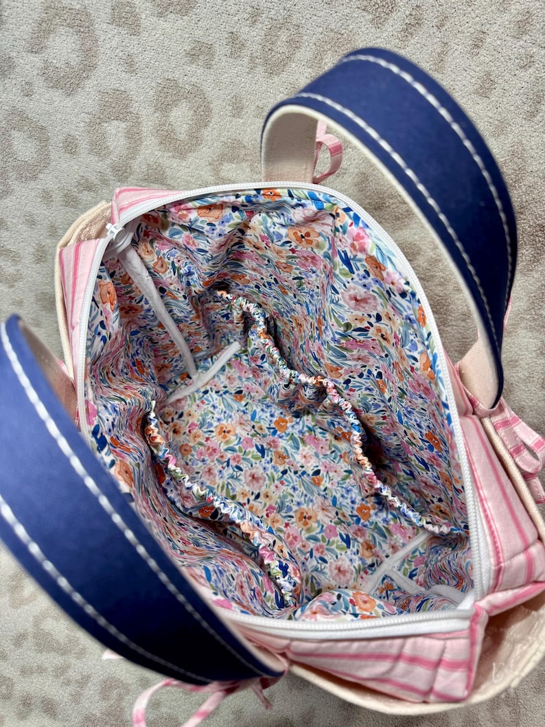 May include: A fabric bag with a pink and white striped exterior and a navy blue handle featuring white stitching. The interior is lined with a floral pattern in shades of pink, orange, blue, and green, and includes an elasticated pocket.