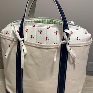 May include: A cream-coloured canvas tote bag featuring two navy blue vertical stripes and matching navy blue handles. The bag has a white quilted insert with a red cherry pattern, a white zip closure, and two small fabric bows tied at the top.