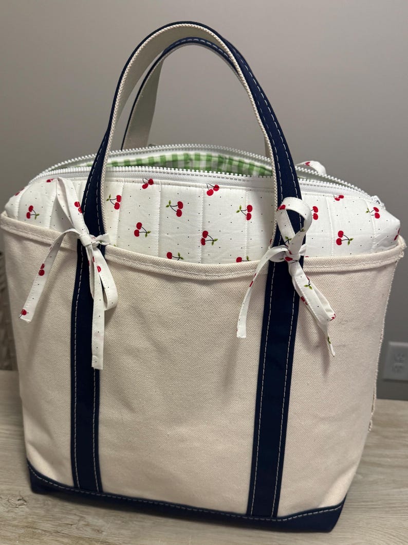 May include: A cream-coloured canvas tote bag featuring two navy blue vertical stripes and matching navy blue handles. The bag has a white quilted insert with a red cherry pattern, a white zip closure, and two small fabric bows tied at the top.
