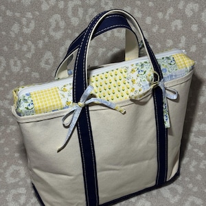 May include: A cream-coloured canvas tote bag featuring dark navy blue handles and a base. The top section includes a zippered closure with a patchwork fabric panel in yellow, white, and blue floral and gingham patterns, accented by small fabric ties.
