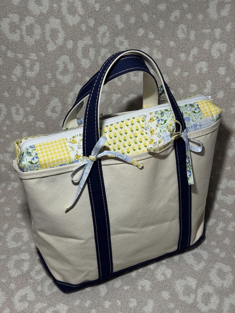 May include: A cream-coloured canvas tote bag featuring dark navy blue handles and a base. The top section includes a zippered closure with a patchwork fabric panel in yellow, white, and blue floral and gingham patterns, accented by small fabric ties.