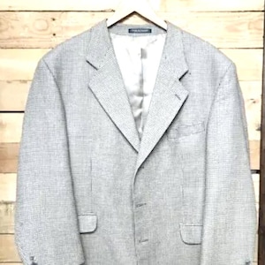 May include: A light gray blazer with a subtle checkered pattern, hanging on a wooden hanger. The jacket has a notched lapel, two front pockets, and buttoned cuffs. The interior lining is a light gray satin.