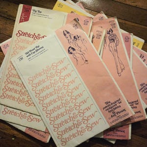May include: A pile of vintage sewing patterns in pink and cream. The patterns feature illustrations of clothing designs and the text "Stretch & Sew". Some patterns include the text "Tab Front Top with Cap Sleeves". The patterns are designed for knit fabrics.