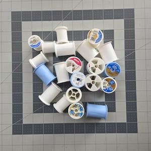 May include: A collection of sewing thread spools in various colors, including white and light blue. The spools are arranged on a gray and black grid surface, showcasing a variety of thread brands and colors.