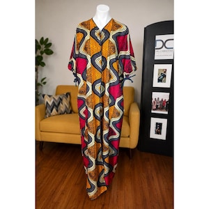 May include: A floor-length kaftan featuring a vibrant geometric wave pattern in shades of mustard yellow, deep magenta, and dark navy blue. The garment has short sleeves with tie closures at the cuffs and a V-neckline, displayed on a white mannequin.