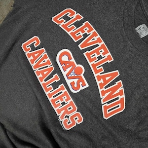May include: Dark gray long-sleeve shirt featuring the words "CLEVELAND CAVALIERS" in orange and white lettering. A "CAVS" logo with a basketball is also visible. The shirt has a crew neck and a tag.