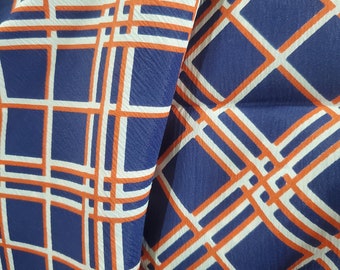 May include: A close-up of a blue fabric with a white and orange geometric pattern. The fabric has a checkered design with overlapping squares and rectangles.