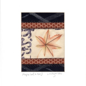 May include: A rectangular textile art piece featuring a central cream panel with a printed orange maple leaf and grey abstract shapes. The panel is bordered by two horizontal bands of brown geometric patterned fabric and dark blue fabric.