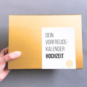Könnte beinhalten: Eine goldfarbene Schachtel mit einem weißen Etikett mit dem Text "DEIN VORFREUDE-KALENDER HOCHZEIT" und einem kleinen gelben Smiley. Die Schachtel wird von einer Hand gehalten, was darauf hindeutet, dass es sich um ein Geschenk oder Paket handelt.