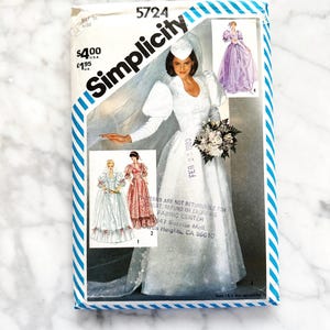 May include: Simplicity sewing pattern number 5724 for a wedding dress. The pattern includes three different views of the dress, all with long sleeves and a fitted bodice. The dress is shown in white with a lace trim. The pattern also includes a veil and a bouquet.