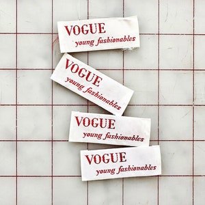 K&ouml;nnte beinhalten: Vier wei&szlig;e Stoff-Etiketten mit rotem Text, der "VOGUE young fashionables" lautet. Die Etiketten sind auf einer wei&szlig;en, karierten Oberfl&auml;che gestapelt.