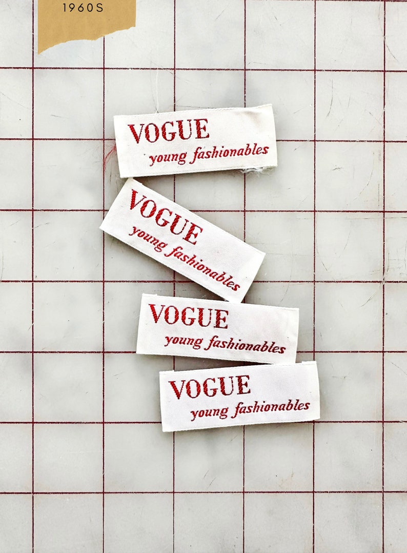 K&ouml;nnte beinhalten: Vier wei&szlig;e Stoff-Etiketten mit rotem Text, der "VOGUE young fashionables" lautet. Die Etiketten sind auf einer wei&szlig;en, karierten Oberfl&auml;che gestapelt.