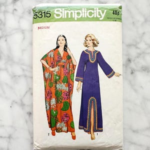 May include: A vintage Simplicity sewing pattern packet featuring two dress designs. The packet is white with green and black text, including "5315" and "Simplicity." The dress illustrations show a long orange and green patterned dress and a long purple dress with gold trim.