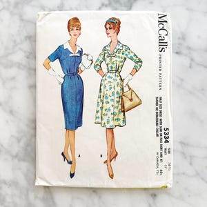 Puede incluir: Patrón de costura impreso vintage de McCall's para un vestido de media talla. El patrón presenta ilustraciones de dos vestidos: un vestido azul con cuello blanco y un vestido con estampado floral. El número de patrón es 5334.