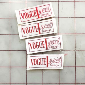 K&ouml;nnte beinhalten: Vier wei&szlig;e Stoff-Etiketten mit rotem Text, der "Vogue Special Design" lautet. Die Etiketten sind vertikal auf einem wei&szlig;en Gitterhintergrund angeordnet.