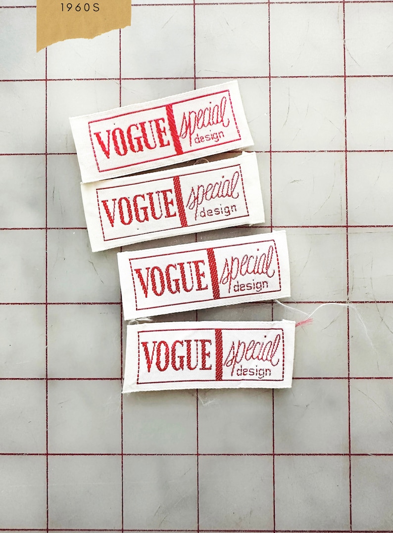 K&ouml;nnte beinhalten: Vier wei&szlig;e Stoff-Etiketten mit rotem Text, der "Vogue Special Design" lautet. Die Etiketten sind vertikal auf einem wei&szlig;en Gitterhintergrund angeordnet.