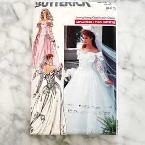 May include: A vintage Butterick sewing pattern featuring illustrations of wedding dresses. The pattern includes a pink off-the-shoulder gown and a white bridal dress with long sleeves. The text on the pattern reads "ADVANCED/PLUS DIFFICILE".