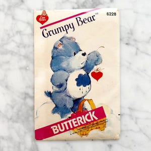May include: A vintage Care Bears sewing pattern featuring Grumpy Bear. The pattern is for a blue bear with a rain cloud on his belly and a red heart in his hand. The pattern number is 6228 and is from the company Butterick.