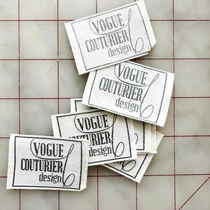K&ouml;nnte beinhalten: Ein Stapel wei&szlig;er Stoff-Etiketten mit schwarzem Text, der "Vogue Couturier design" lautet.