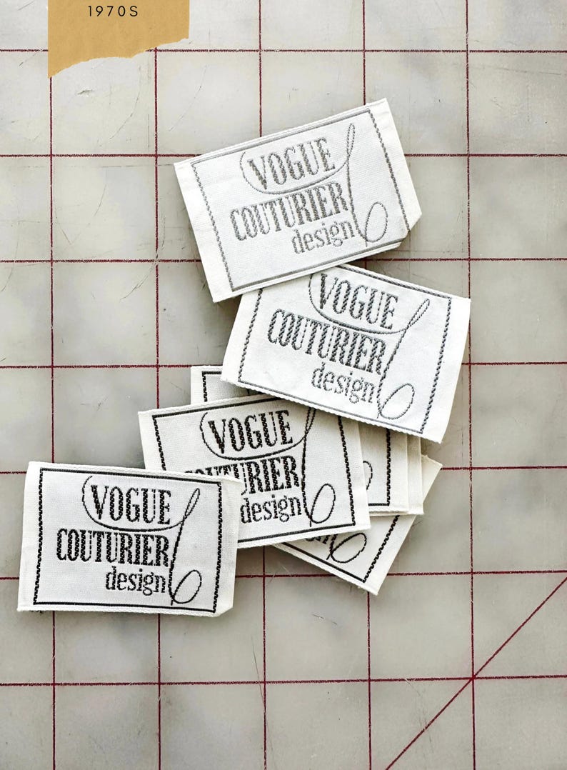 K&ouml;nnte beinhalten: Ein Stapel wei&szlig;er Stoff-Etiketten mit schwarzem Text, der "Vogue Couturier design" lautet.
