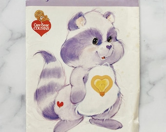May include: A vintage Care Bear Cousins sewing pattern featuring Bright Heart Raccoon. The pattern is purple and white with a picture of the character. The pattern number is 3380.