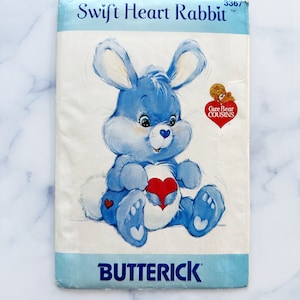 May include: A vintage sewing pattern for a Care Bear Cousins plush toy. The pattern is for a blue rabbit with a red heart on its chest. The pattern is titled "Swift Heart Rabbit" and is number 3367. The pattern is from the company Butterick.