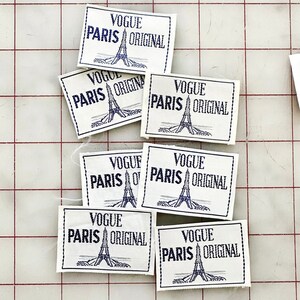 K&ouml;nnte beinhalten: Eine Sammlung von sieben Vintage-Stoffetiketten aus wei&szlig;em Stoff mit blauer Stickerei. Jedes Etikett tr&auml;gt den Text "Vogue Paris Original" und ein stilisiertes Bild des Eiffelturms.