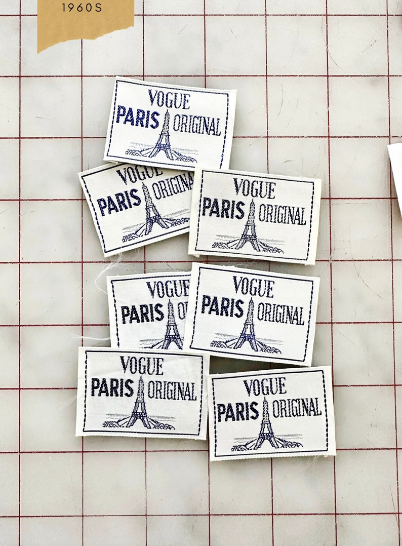 K&ouml;nnte beinhalten: Eine Sammlung von sieben Vintage-Stoffetiketten aus wei&szlig;em Stoff mit blauer Stickerei. Jedes Etikett tr&auml;gt den Text "Vogue Paris Original" und ein stilisiertes Bild des Eiffelturms.