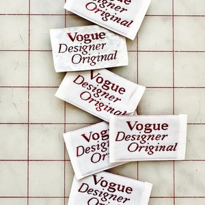K&ouml;nnte beinhalten: F&uuml;nf wei&szlig;e Stoff-Etiketten mit rotem Text, der "Vogue Designer Original" lautet. Die Etiketten sind &uuml;bereinander auf einem wei&szlig;en Gitterhintergrund gestapelt.