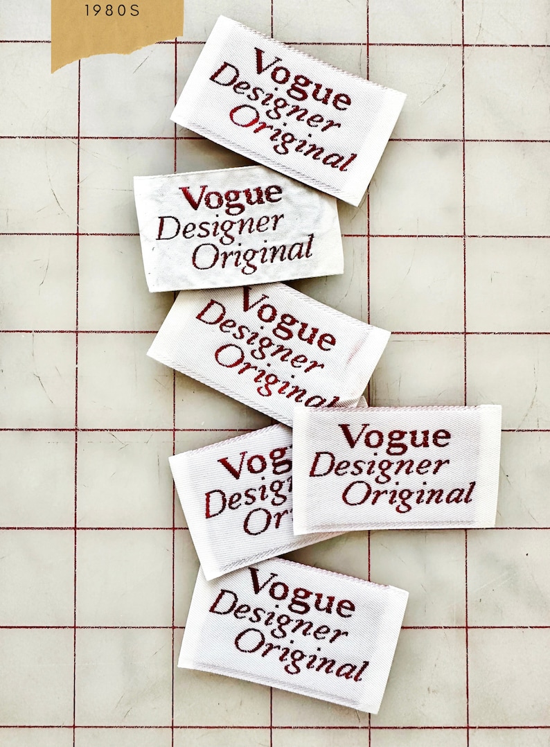K&ouml;nnte beinhalten: F&uuml;nf wei&szlig;e Stoff-Etiketten mit rotem Text, der "Vogue Designer Original" lautet. Die Etiketten sind &uuml;bereinander auf einem wei&szlig;en Gitterhintergrund gestapelt.