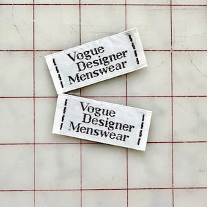 K&ouml;nnte beinhalten: Zwei wei&szlig;e Stoff-Etiketten mit schwarzem Text, der "Vogue Designer Menswear" lautet.
