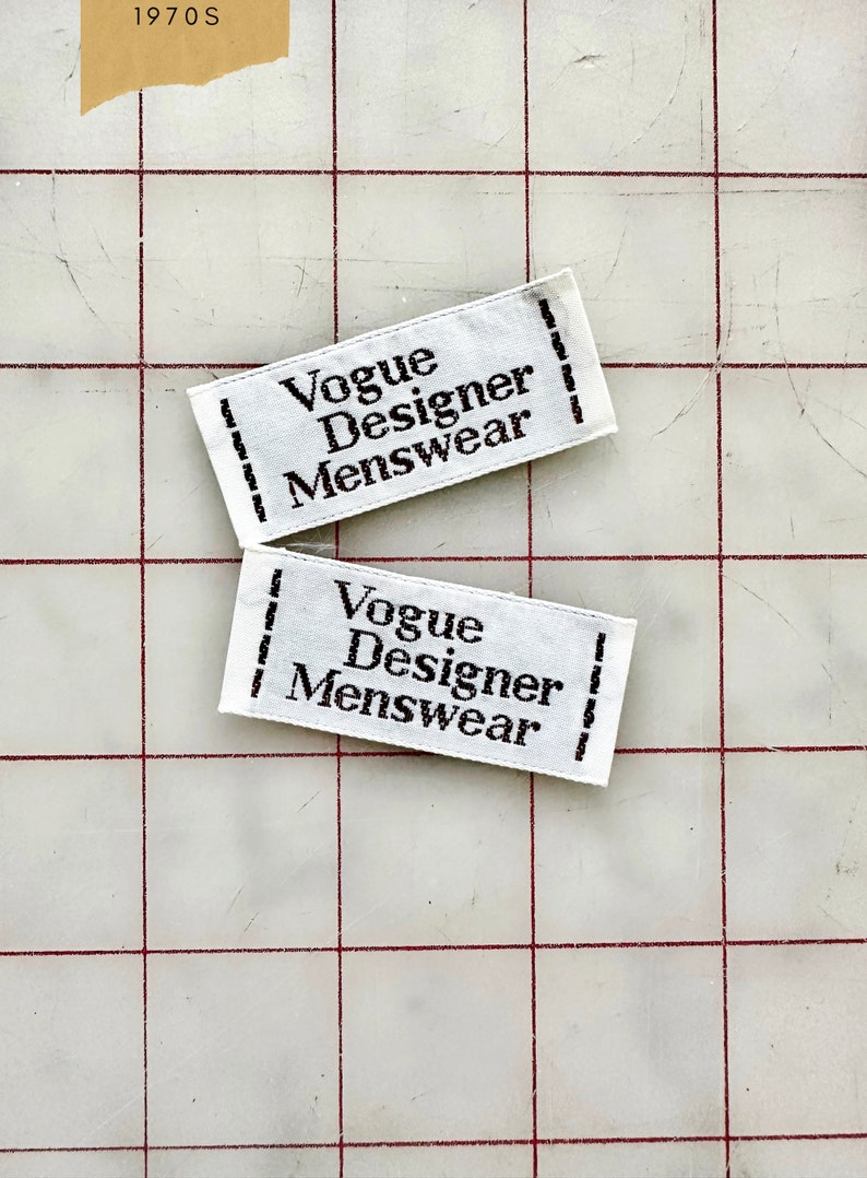 K&ouml;nnte beinhalten: Zwei wei&szlig;e Stoff-Etiketten mit schwarzem Text, der "Vogue Designer Menswear" lautet.