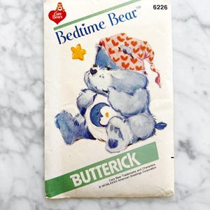 May include: A vintage sewing pattern for a Care Bear bedtime bear. The pattern is from Butterick and features a blue bear wearing a pink and white heart-patterned hat. The pattern number is 6226.