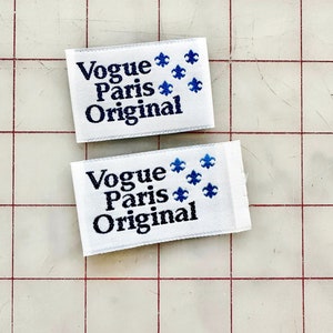 K&ouml;nnte beinhalten: Zwei wei&szlig;e Stoff-Etiketten mit blauen gestickten Fleur-de-lis und schwarzem Text, der "Vogue Paris Original" lautet.