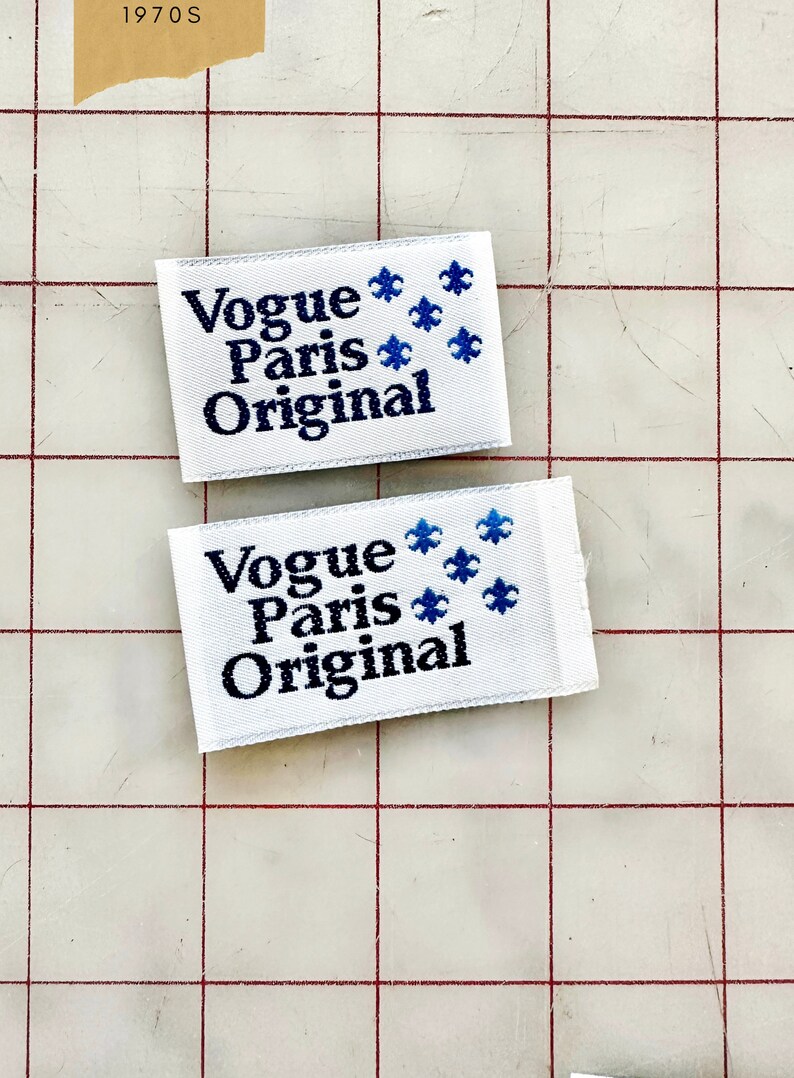 K&ouml;nnte beinhalten: Zwei wei&szlig;e Stoff-Etiketten mit blauen gestickten Fleur-de-lis und schwarzem Text, der "Vogue Paris Original" lautet.