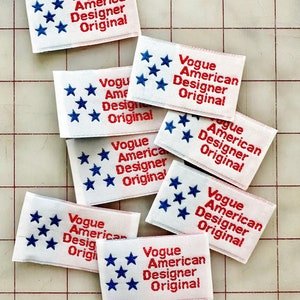 K&ouml;nnte beinhalten: Nahaufnahme von mehreren wei&szlig;en Stoff-Etiketten mit roten und blauen Stickereien. Die Etiketten tragen die Aufschrift "Vogue American Designer Original" und zeigen f&uuml;nf blaue Sterne.