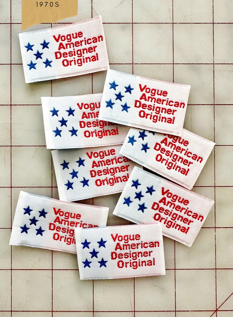 K&ouml;nnte beinhalten: Nahaufnahme von mehreren wei&szlig;en Stoff-Etiketten mit roten und blauen Stickereien. Die Etiketten tragen die Aufschrift "Vogue American Designer Original" und zeigen f&uuml;nf blaue Sterne.