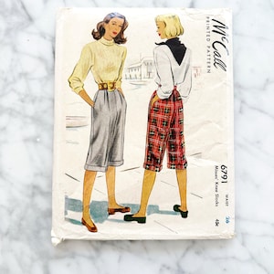 以下が含まれることがあります： ミセス・ニー・スラックスのヴィンテージMcCall'sプリントパターン。2人の女性がスタイリッシュな服装で描かれています。表紙には、グレーのショートパンツを着た女性と、赤いチェックのショートパンツを着た女性が描かれています。パターン番号は6791で、ウエストサイズは26です。