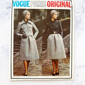 May include: Vogue Paris Original pattern cover featuring two models wearing gray outfits. One model wears a jacket with a collar and a skirt, while the other wears a turtleneck sweater and a skirt. Both models are wearing hats and black shoes. The text "Vogue Paris Original" is printed in red, blue, and black letters.
