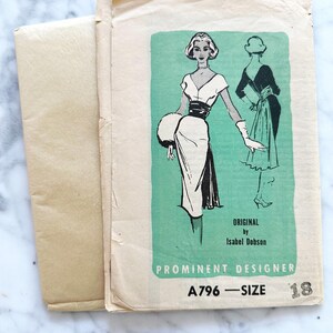 May include: Vintage sewing pattern featuring a dress design. The pattern includes a drawing of a woman in a white dress with a black belt and a matching fur muff. The text reads "Original by Isabel Dobson" and "Prominent Designer".