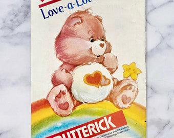 May include: A vintage sewing pattern featuring Love-a-Lot Bear from the Care Bears. The pattern is for a pink bear with a heart on its chest, sitting on a rainbow. The pattern is from the company Butterick and is number 305.