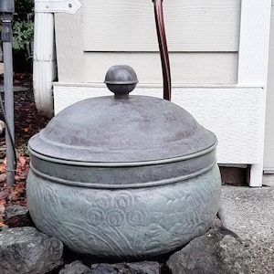 May include: A round, gray metal garden container with a domed lid and a spherical knob handle. The exterior features a relief pattern of floral and leaf motifs. A dark red hose extends upward from behind the lid of the container.