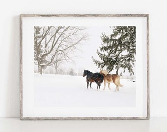 May include: A framed print featuring three horses in a snowy landscape. One horse is black, and the other two are light brown. The scene includes bare trees and a large evergreen tree, with snow falling.