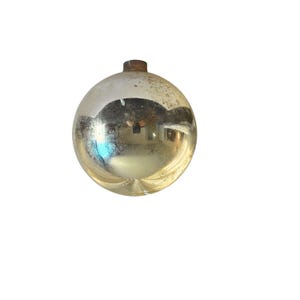 May include: A vintage, round Christmas ornament with a tarnished, reflective surface. The ornament has a metallic gold color with areas of discoloration and a small, rusty-colored metal cap at the top. The surface reflects the surrounding environment.
